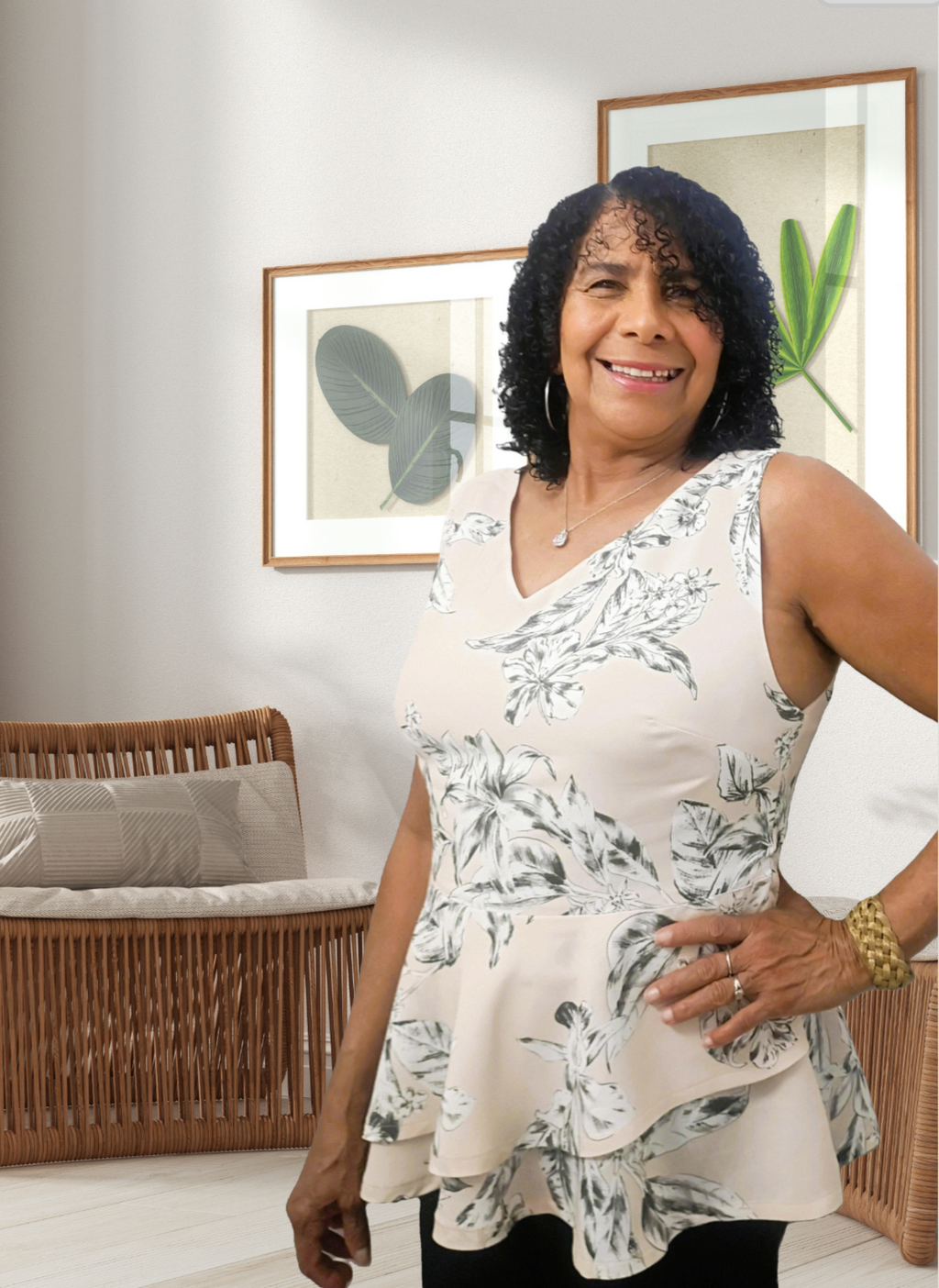 A front-facing lifestyle photo of a smiling woman modeling the sleeveless V-neck peplum blouse. The lightweight top features a delicate black botanical floral print over a light beige background, highlighting the flowy, layered asymmetrical hem. She is standing in a brightly lit, beautifully styled modern room.