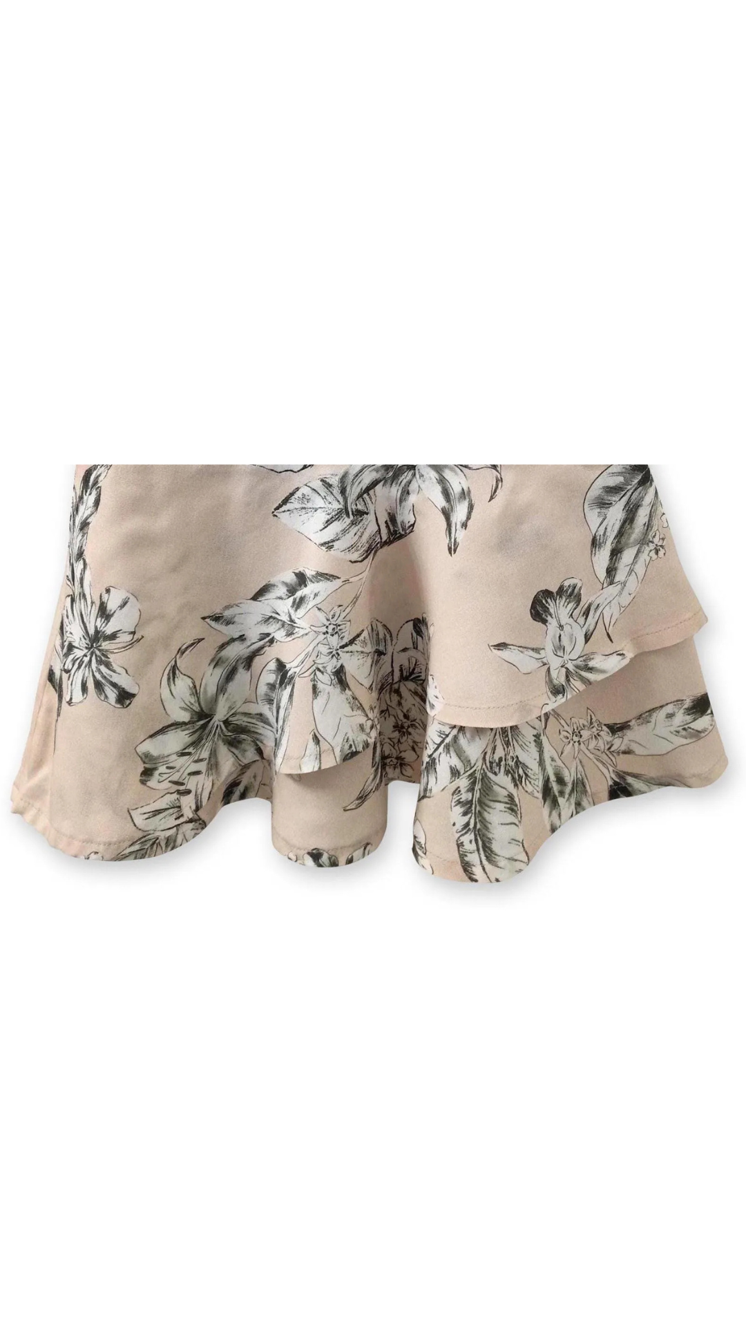 A close-up macro photograph highlighting the delicate black botanical floral pattern printed on the soft, lightweight beige Ecovero viscose fabric. The shot focuses on the fine details of the illustrated plan motifs, surrounded by a warm, naturally lit background featuring leafy shadows.